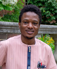 A portrait of a smiling man wearing a traditional pink outfit, seated outdoors with greenery in the background.