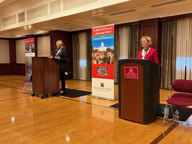 Two speakers at a podium during a discussion event at Miami University, with banners showcasing the event theme in the background.