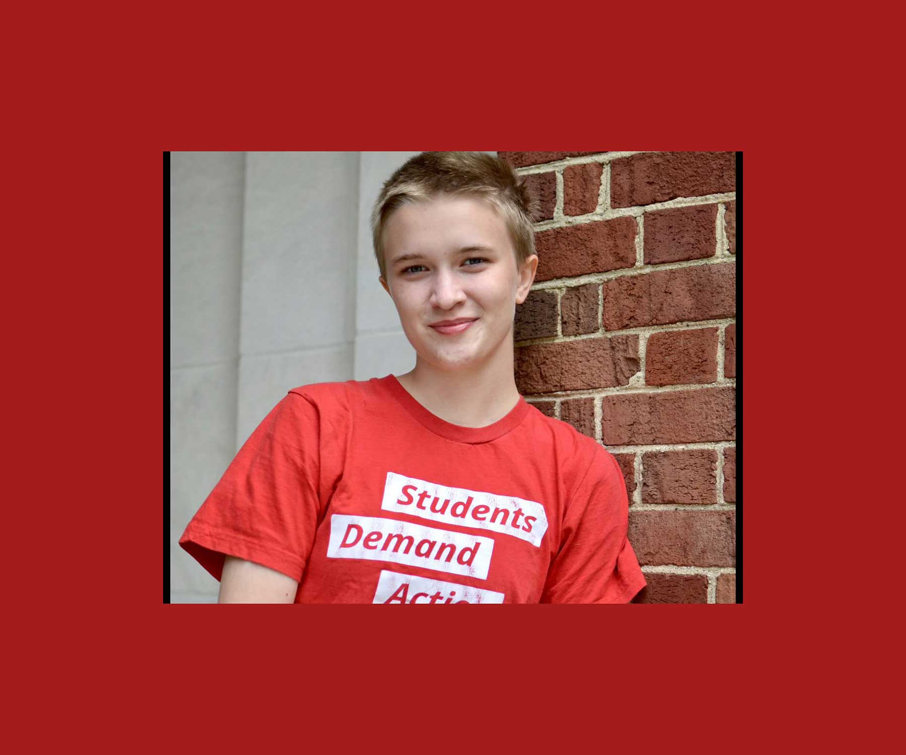 Student Peren Tiemann stands in front of a brick wall. They have short blond hair and are wearing a read t-shirt which reads students demand action