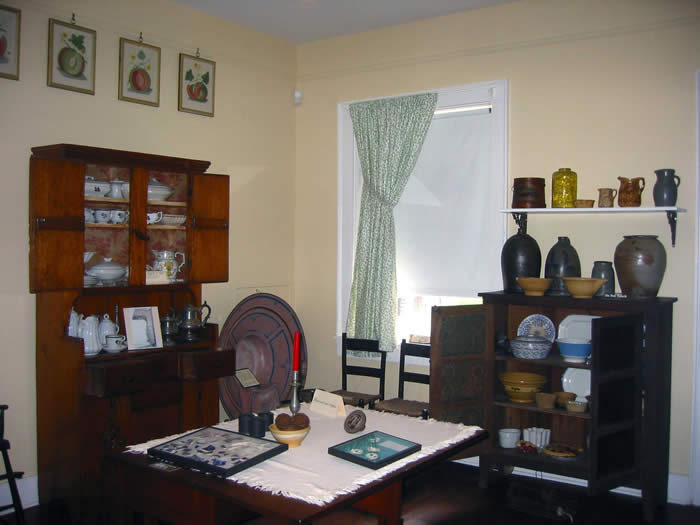 A room with kitchen furniture, including a table set for a meal, ladderback chairs, a tall cupboard and a short pie safe cupboard displaying dishes and cookware