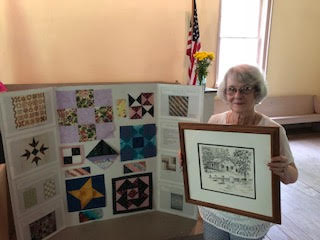Sue Jones holding a framed sketch of a pioneer building near a trifold display of quilt blocks