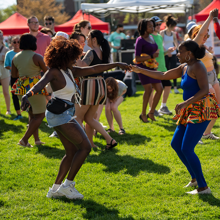 Candid photo of two Black women dancing on a large grassy lawn with crowds of people and red tents surrounding them
