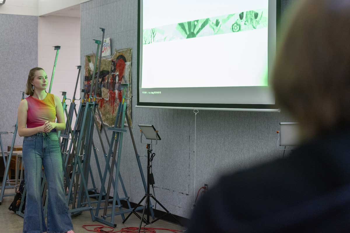 Artist talking to an audience with artwork projected on the screen beside her.