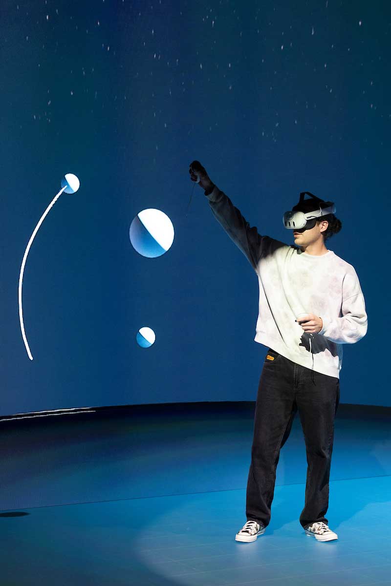 Person in VR headset reaches toward floating virtual planets in a dark, immersive environment.