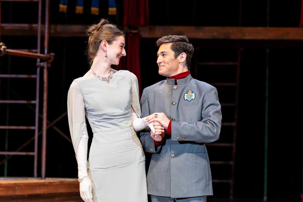 Two actors perform on stage in elegant period costumes; the woman wears a long, light-colored gown with gloves and a necklace, while the man wears a gray military-style uniform with a crest. They smile at each other, holding hands in a romantic gesture.