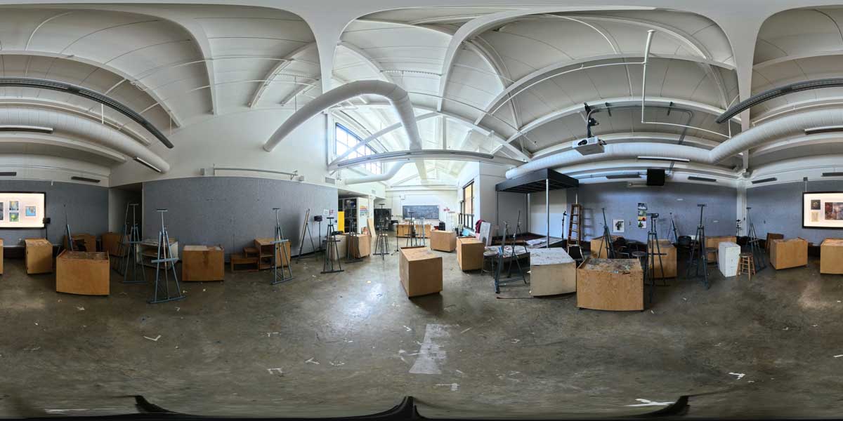 360-degree view of a large art studio with high arched ceilings, exposed ducts, easels, work tables, and stools. Artwork hangs on the walls, and the concrete floor has scattered supplies. Large windows provide natural light.