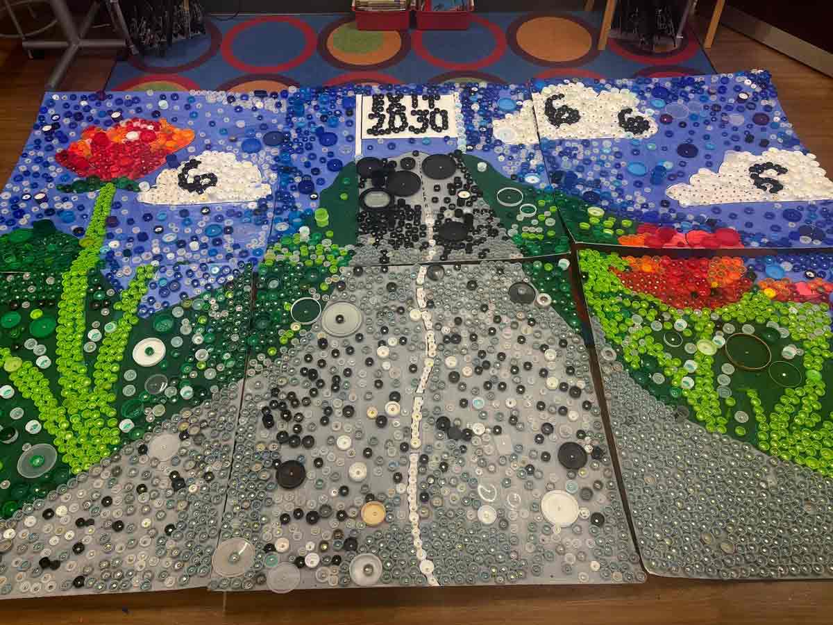 six panels placed together decorated with buttons creating an art piece depicting flowers along a road with clouds in the sky and a banner at the end of the road that says Exit 2030