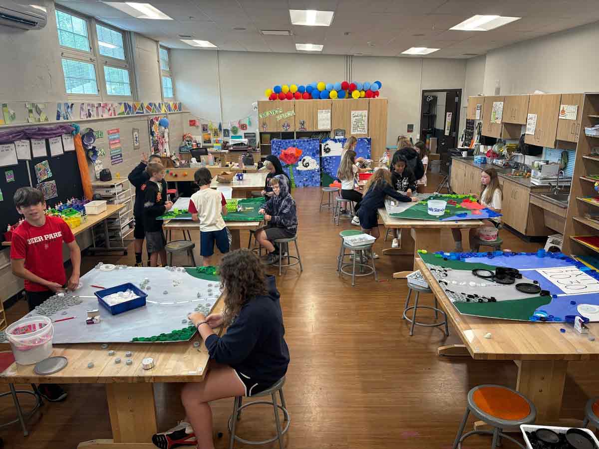 elementary students work in teams to create six panels placed together decorated with various materials creating an art piece depicting flowers along a road with clouds in the sky and a banner at the end of the road that says Exit 2030