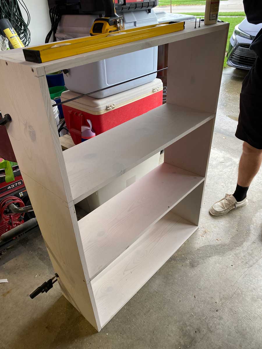 The nearly assembled white wooden shelf stands upright in a garage, with tools and supplies nearby.