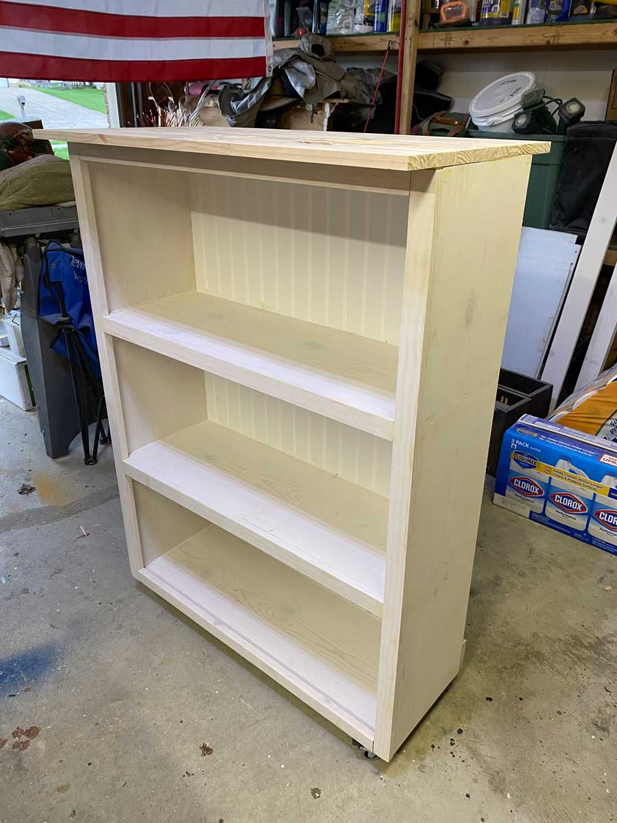 The fully built wooden shelf, unpainted but complete with three interior shelves and a decorative top, positioned in a workshop.