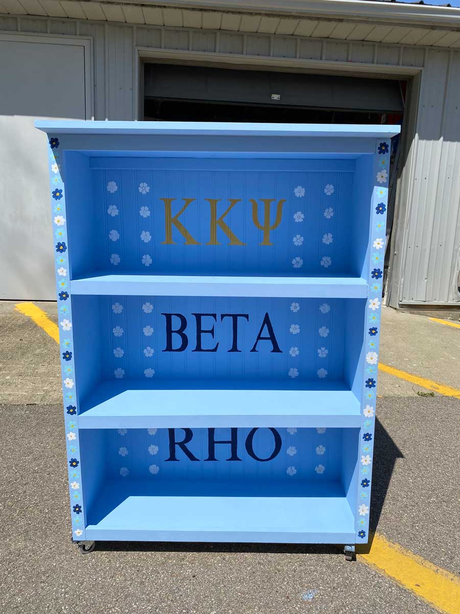 The completed shelf, now painted bright blue with decorative white and dark blue flowers, features the Greek letters Kappa Kappa Psi and the words Beta Rho on the back panel.