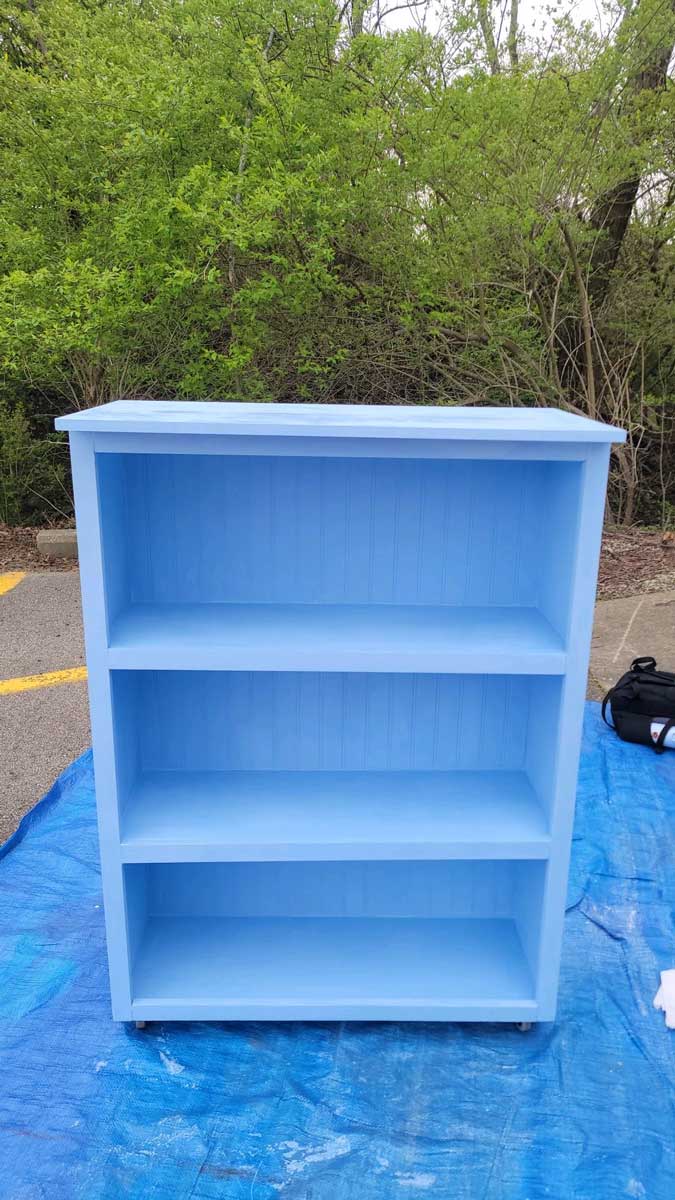 The freshly painted blue shelf, without lettering or decoration, is drying outdoors on a tarp near a wooded area.