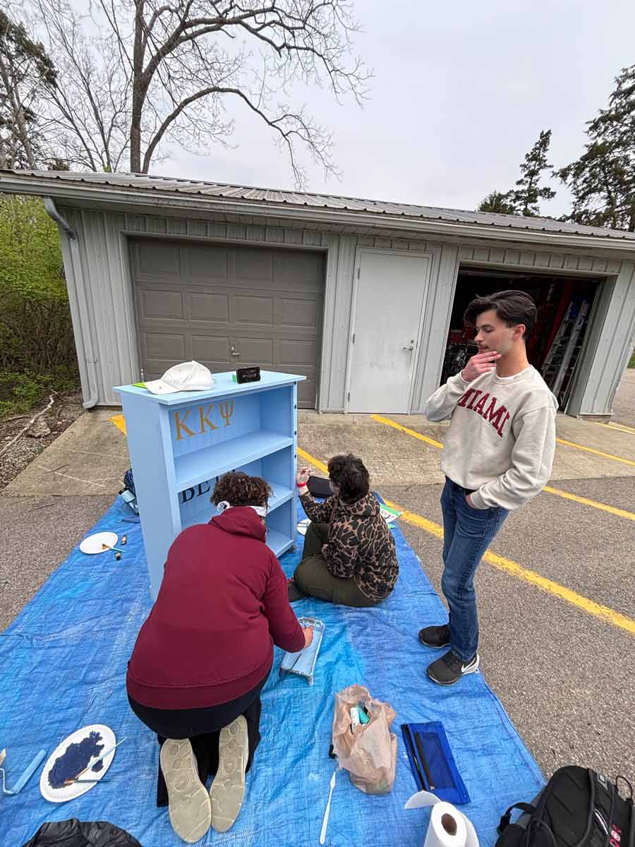Three students crouch and sit while painting details and lettering on the blue shelf outside, surrounded by supplies and paint trays.