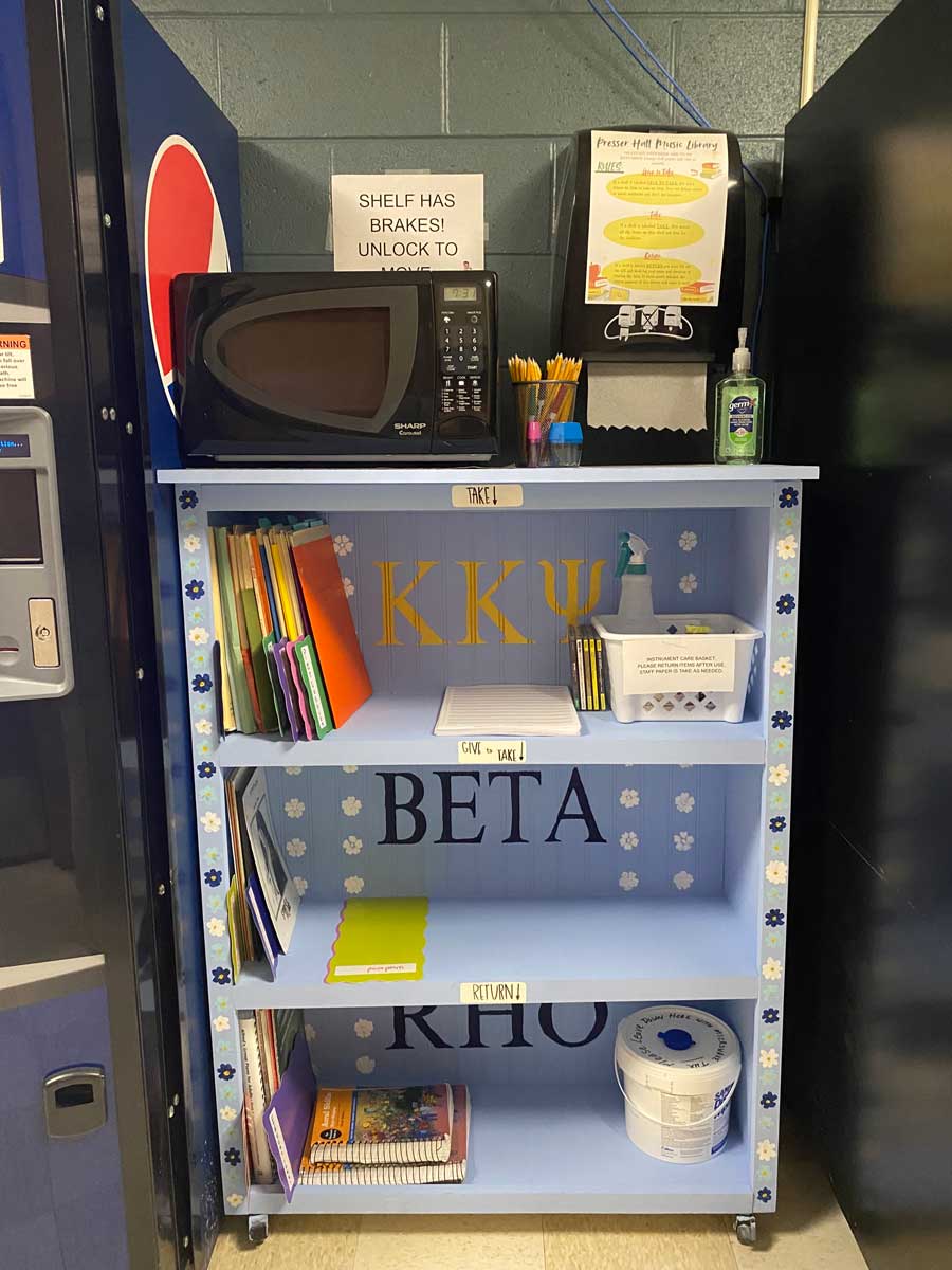 The stocked music library shelf in use, filled with sheet music, educational books, supplies, and a microwave, with signs indicating Take, Give and Take, and Return sections.