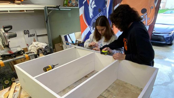Two students in a garage workshop carefully measure and assemble a wooden structure as part of the music library shelf construction.