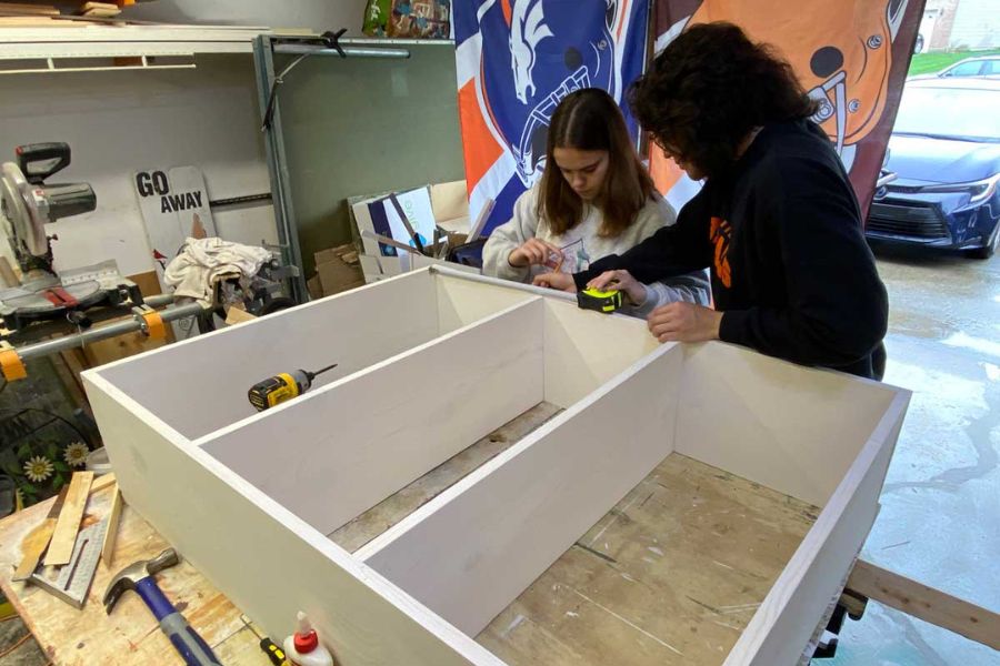 Two students in a garage workshop carefully measure and assemble a wooden structure as part of the music library shelf construction.