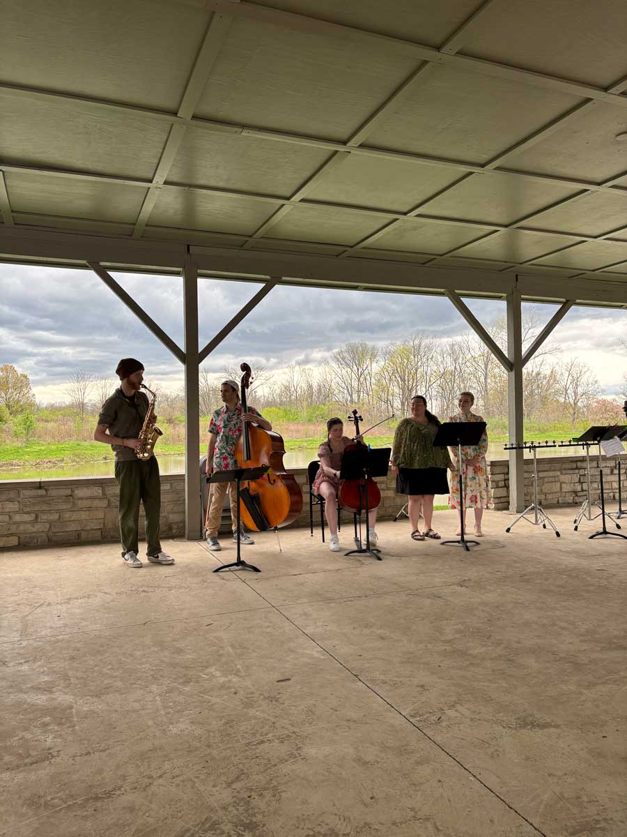 A small ensemble of five musicians performing under a covered outdoor pavilion. Instruments include a saxophone, upright bass, cello, and vocals, with music stands in front of them. The background shows a grassy field and overcast skies.