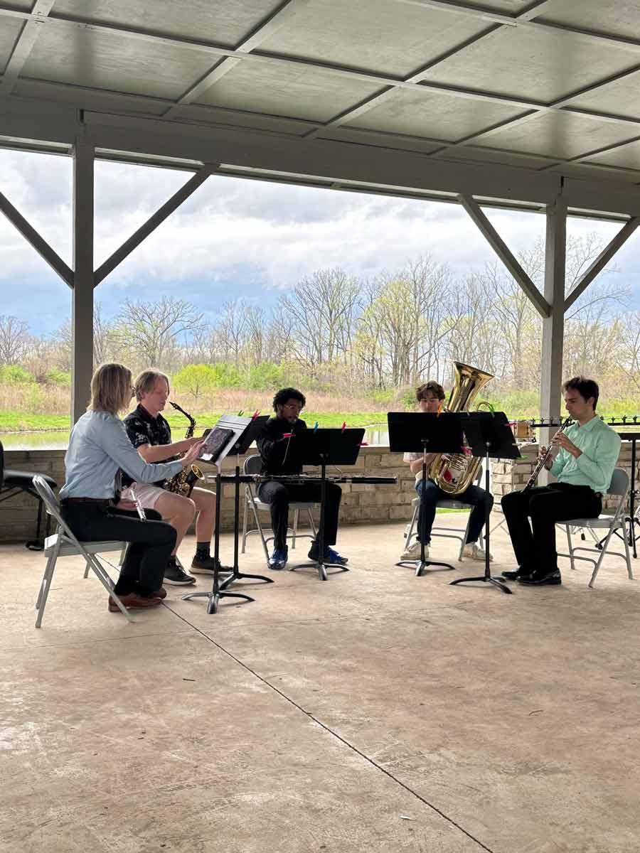 A different group of five musicians performing, including a clarinet, saxophone, euphonium, and other woodwind and brass instruments. They are seated in a semicircle with music stands, also under the pavilion.
