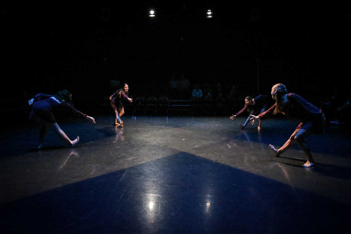 Four dancers perform expressive, low movements in a dark theater