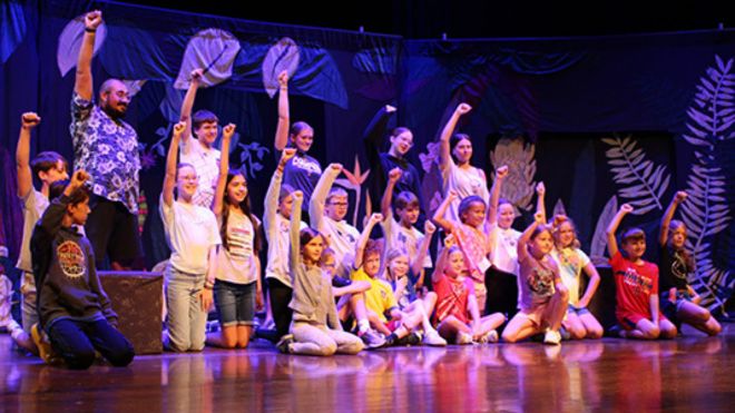 Students gathered on a theater stage, some standing  and some kneeling, each with their right first raised in the air at the end of a production.
