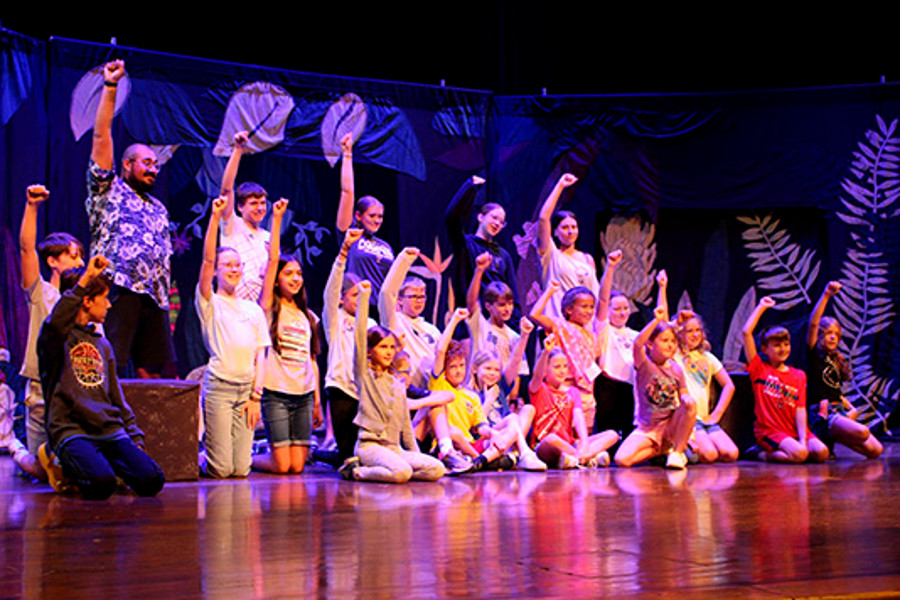 Students gathered on a theater stage, some standing  and some kneeling, each with their right first raised in the air at the end of a production.