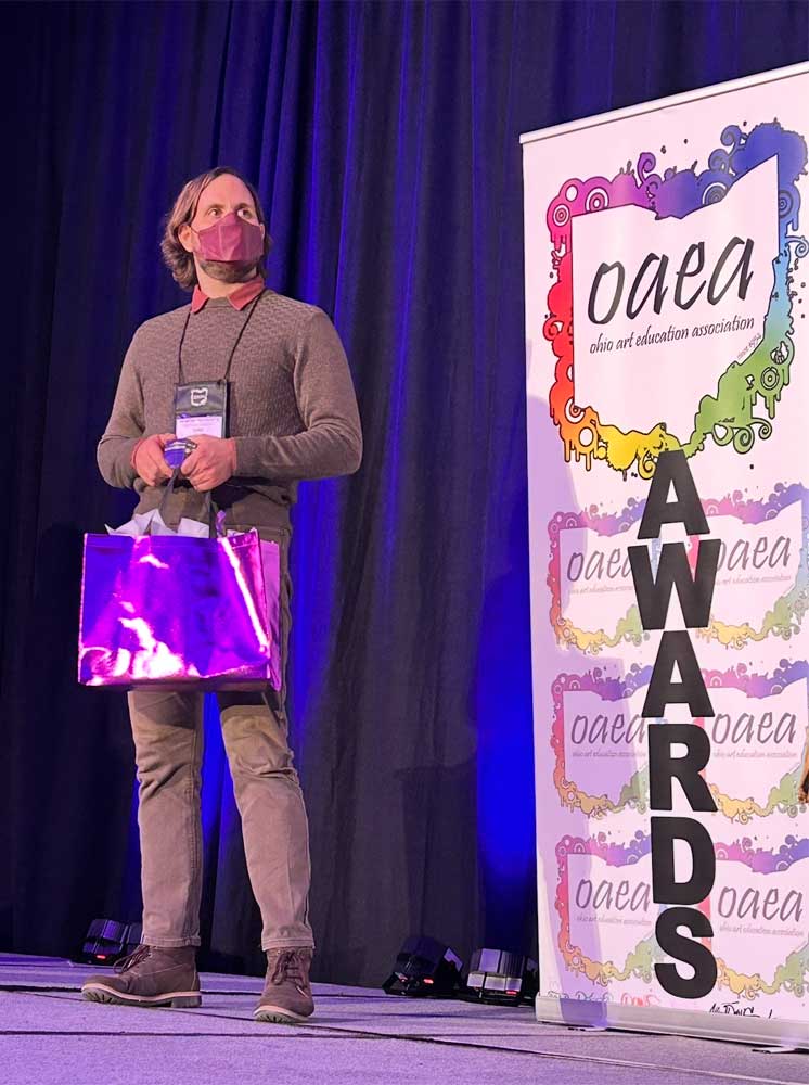 Miami University faculty member Luke Meeken stands onstage with a pink gift bag during the 2025 OAEA Awards presentation.