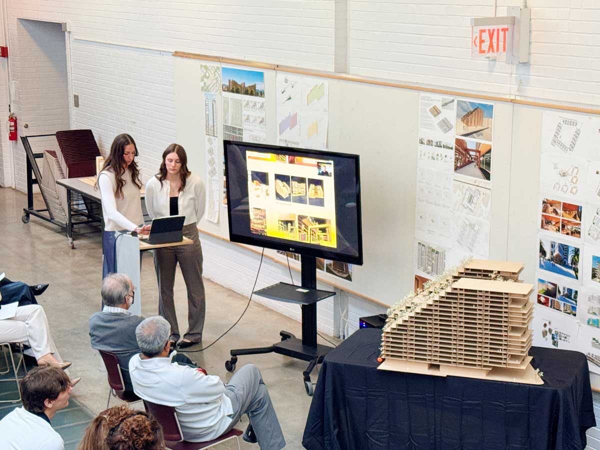 Amber Tretter and Zoe Cowles give a presentation beside an architectural model, with project boards and a screen, in front of an audience.