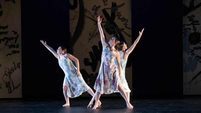 Three dancers perform a contemporary dance onstage, each wearing flowing sleeveless dresses and extending their arms upward against a dark backdrop with abstract imagery.