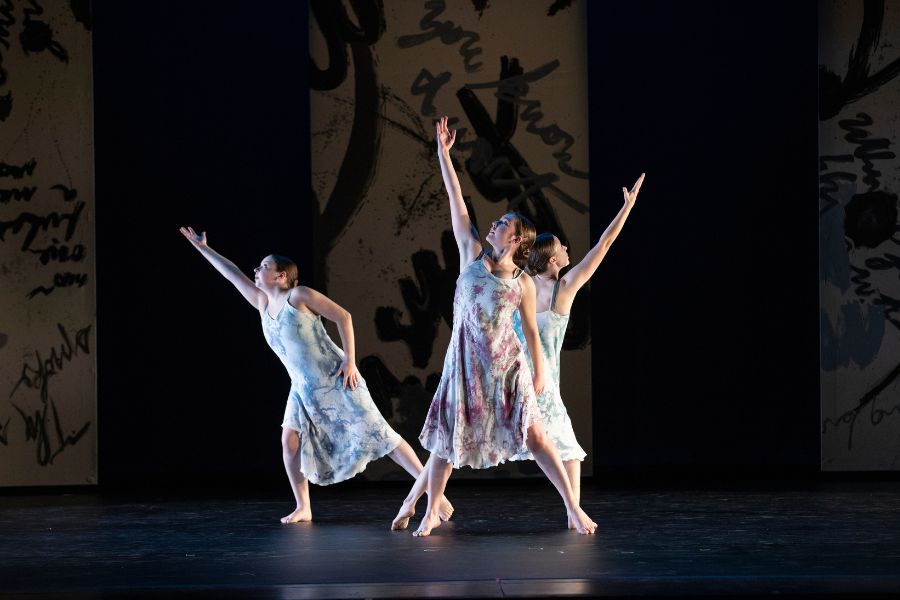 Three dancers perform a contemporary dance onstage, each wearing flowing sleeveless dresses and extending their arms upward against a dark backdrop with abstract imagery.