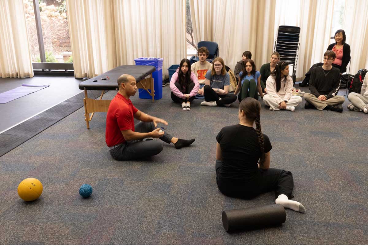 Dr. Jason Gross, wearing a red collared shirt, leads Miami University students in a whole-body musicianship class, seated in a circle with therapy balls and foam rollers in a studio classroom.