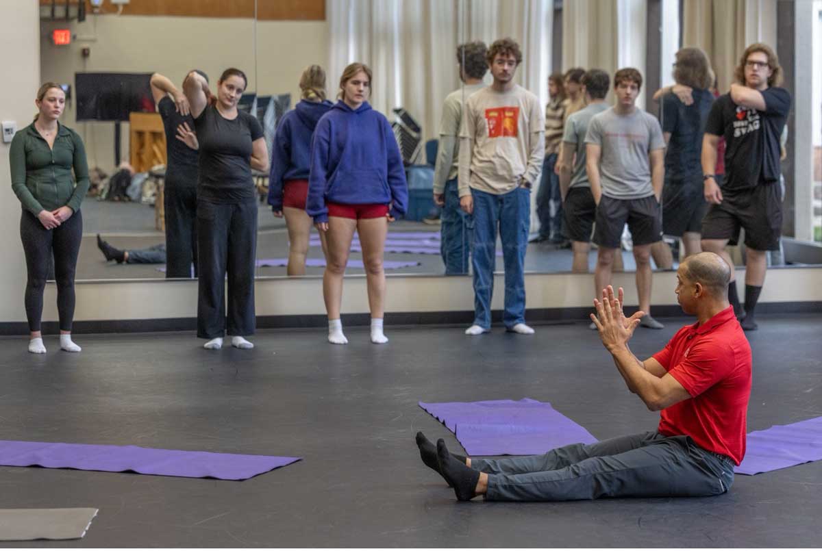 Dr. Jason Gross, wearing a red collared shirt, demonstrates posture and movement techniques as Miami University music students stand on yoga mats in a mirrored studio classroom.