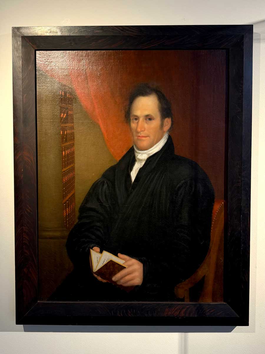 Portrait painting of Miami University’s first president, Robert Hamilton Bishop, seated in a dark robe and holding a book, with a red curtain and tall bookshelf behind him.
