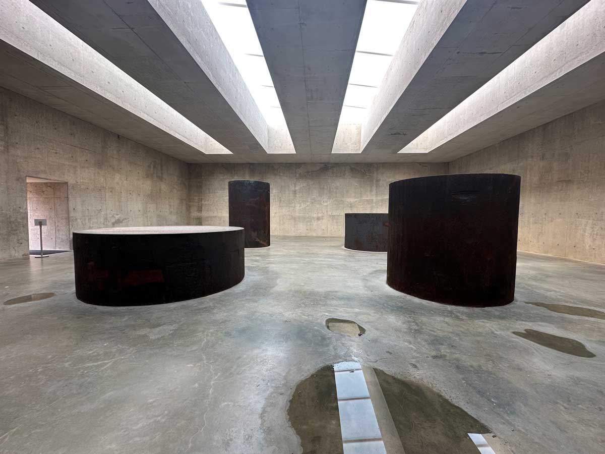 Minimalist gallery with large cylindrical sculptures and skylights titled Four Rounds Equal Weight, Unequal Measure by Richard Serra at Glenstone Museum.