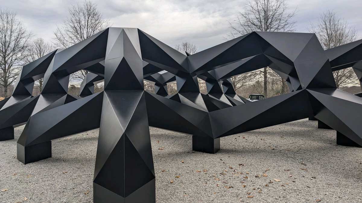 Large black geometric outdoor sculpture installation titled Smug by Tony Smith at Glenstone Museum surrounded by trees.