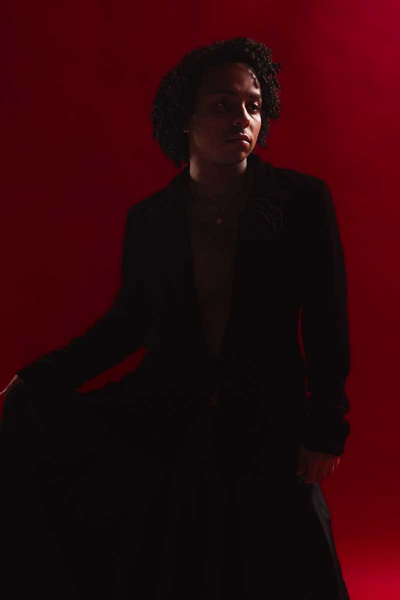 Model in a black suit seated against a red backdrop, lit dramatically from the side.