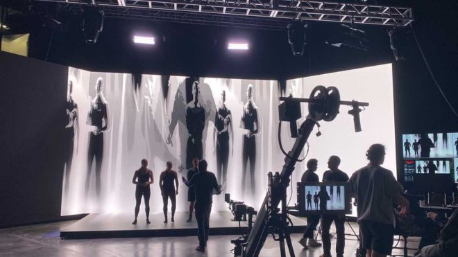 Studio production with performers, crew, and a large digital backdrop showing black-and-white figures.