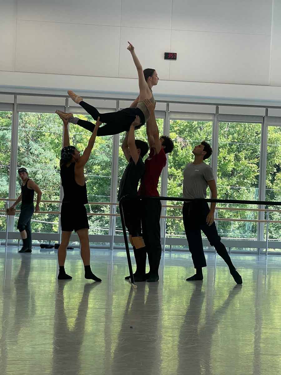 Cincinnati Ballet dancers rehearse a group lift, supporting a dancer with legs extended behind and one arm reaching upward.