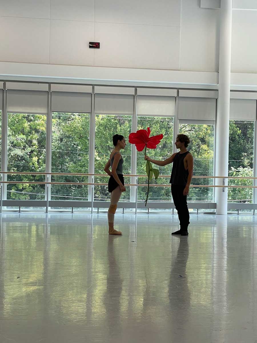 Two Cincinnati Ballet dancers face each other in a sunlit studio, one holding an oversized red flower prop between them.