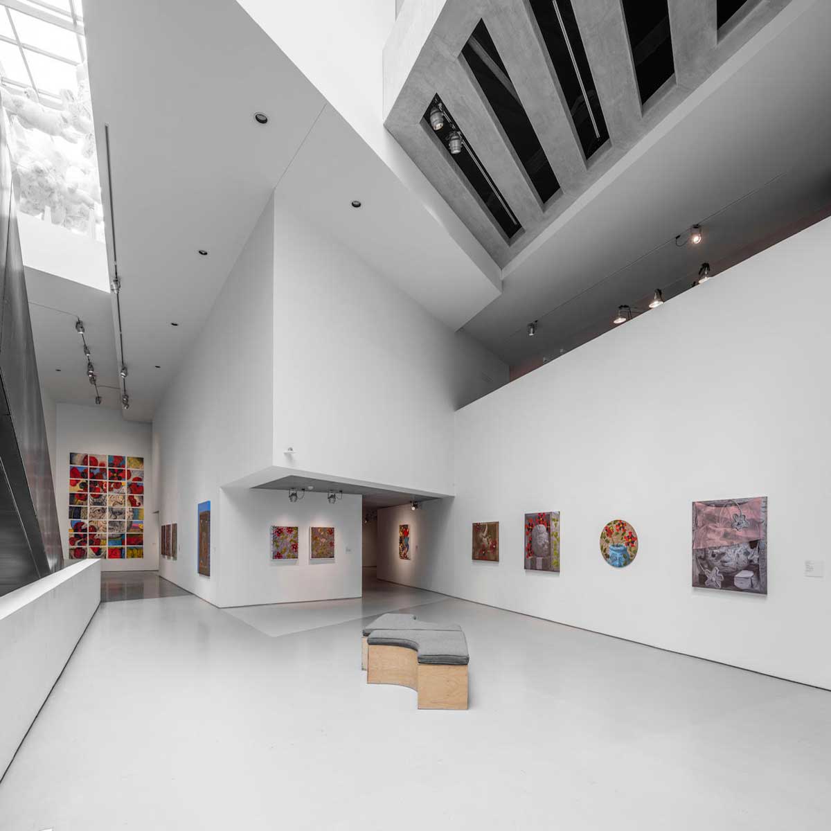 Colorful paintings line a high-ceilinged gallery, with curved benches in the center and a tall, gridded painting on the far wall.