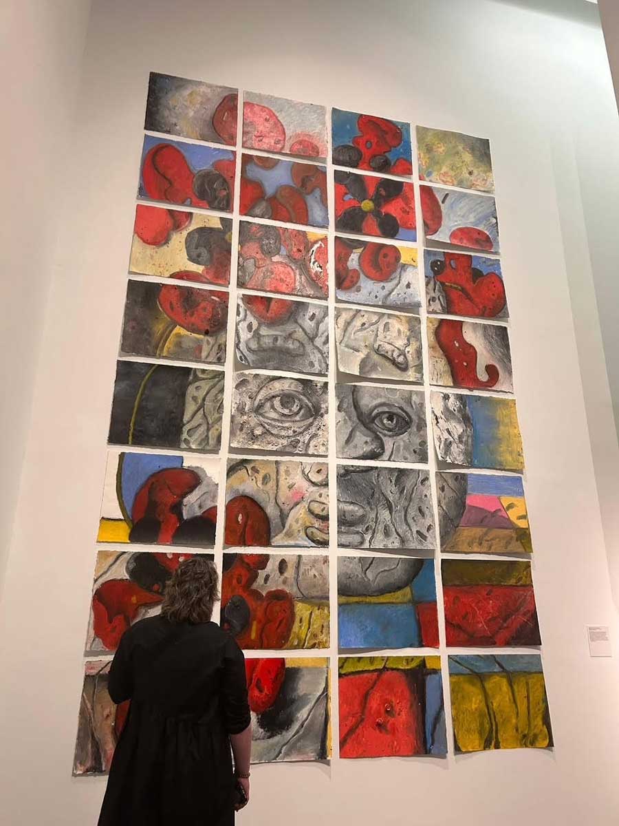 A visitor looks up at Michael Stillion’s large grid of painted panels forming a gray face surrounded by red flowers.