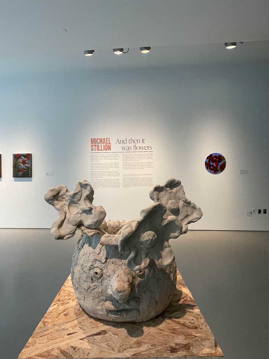 Michael Stillion’s rough-textured gray vessel has a sculpted face and large flowerlike forms rising from its rim atop a wooden pedestal.