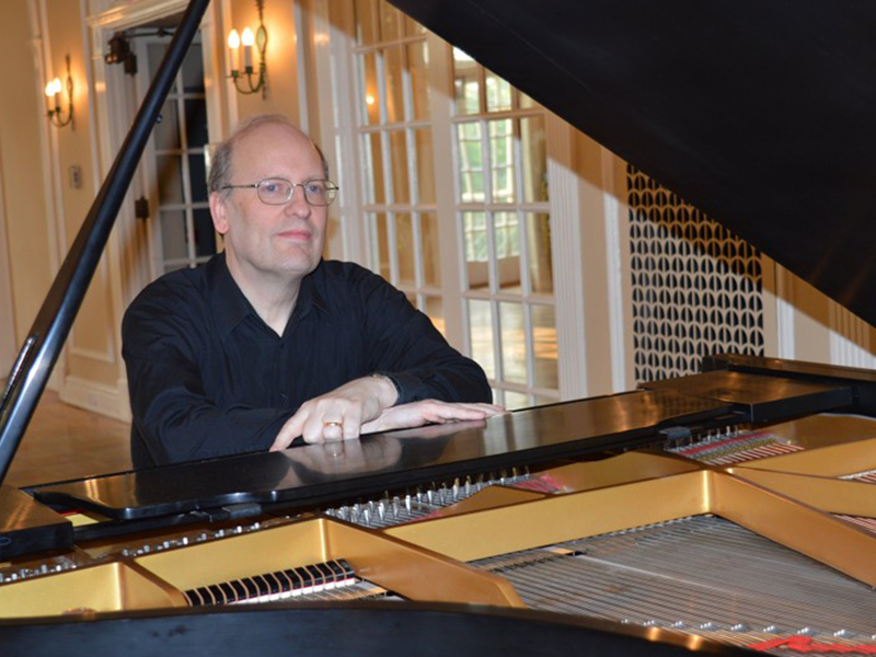 A white man with grey hair and glasses sits at a grand piano playing with passion