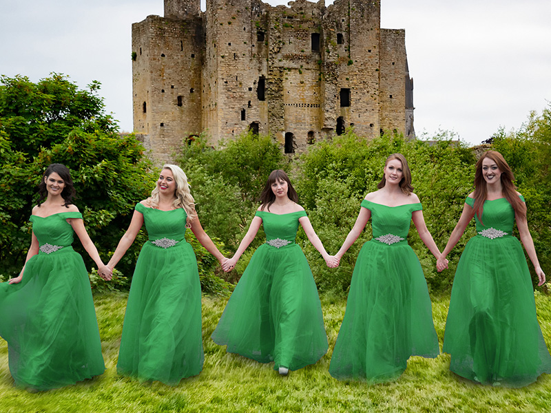 A group of women wearing long flowing green dresses stand in front of the ruins of an Irish Castle