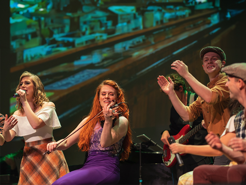 A woman plays a violin as people around her clap and sign along in an irish pub type setting