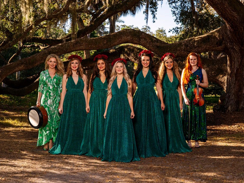 five women dressed in long flowing gowns stand in a forest setting 