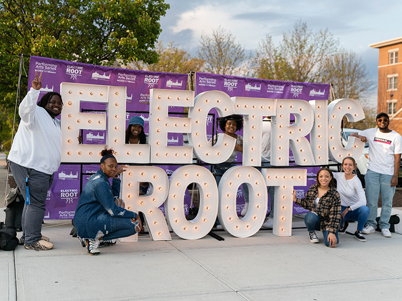 A group of students stand beside giant light up letters that spell Electric Root