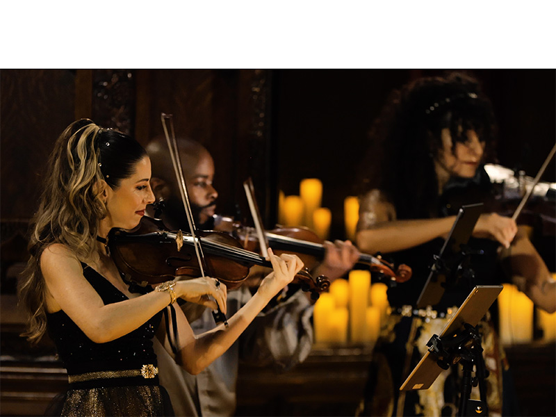 Three performers playing violins and a viola play on a stage surrounded by lit candles 