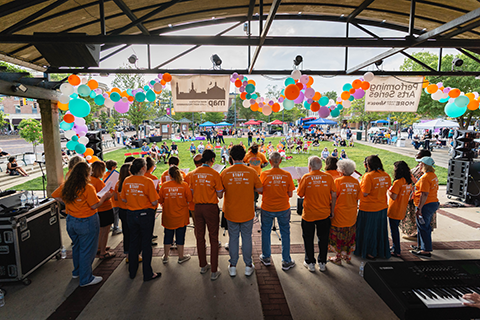 A photo of several college student wearing bright aqua t-shirts with the Electric Root Festival logo standing at a table helping children with an arts project