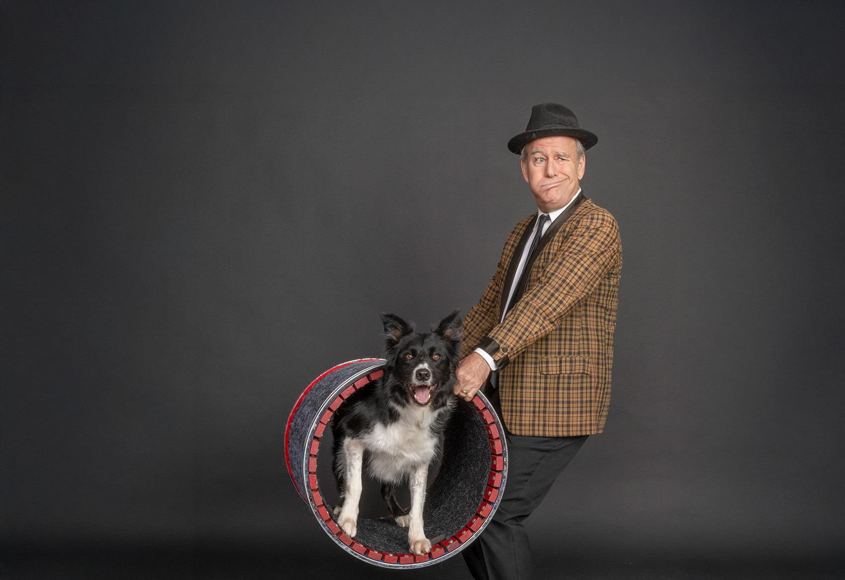 Photo of an older man wearing a comedically patterned suit jacket and fedora hat holds a open ended barrel with a black and white dog of mixd breed standing inside the barrel looking at the camera.