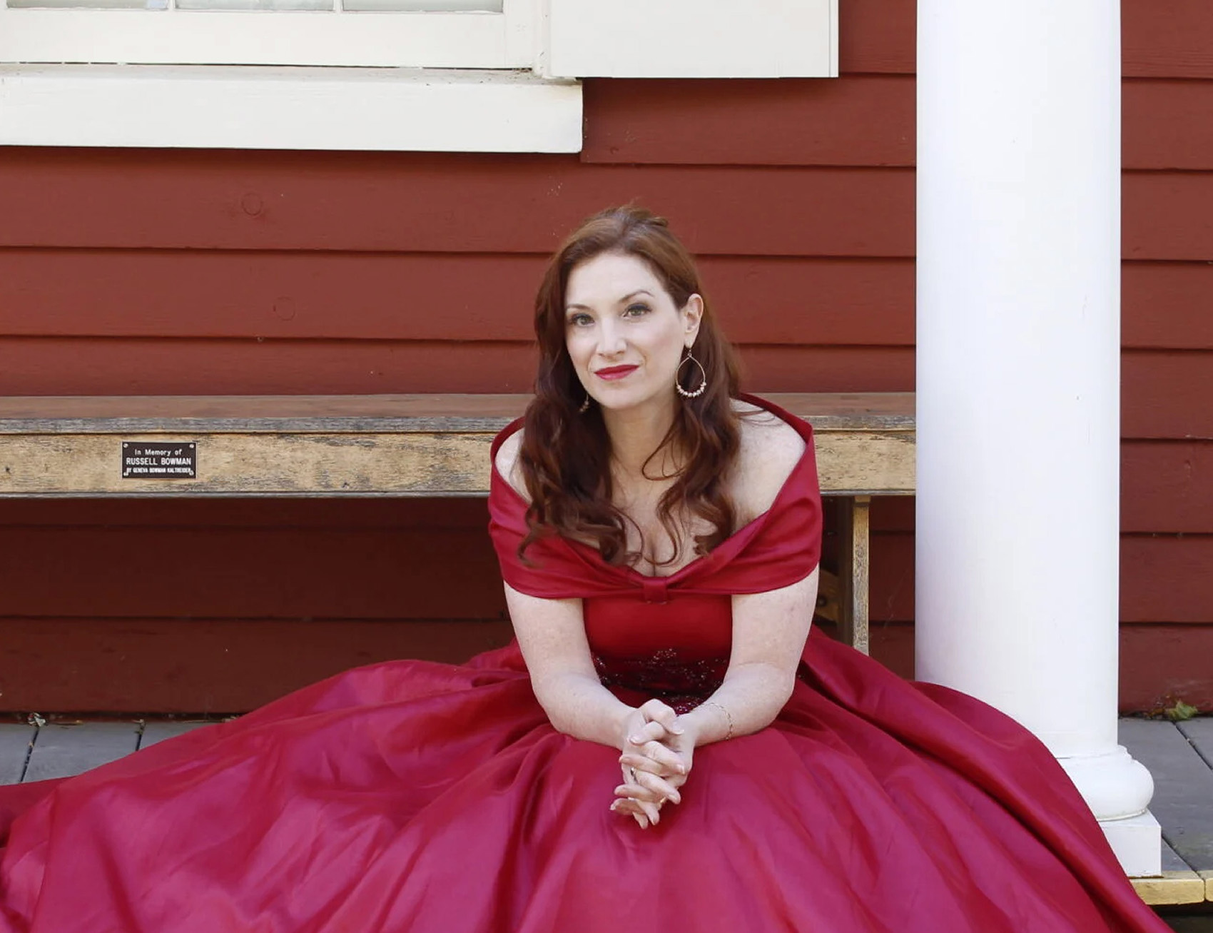 Robin Spielberg a woman with long wavy brown hair sits on the steps of a wooden porch in a bright red formal dress with a large skirt that is spread out around her